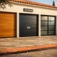 Fachada mediterránea con tres puertas de garaje: madera de roble, acero gris y vidrio, bajo luz cálida. Cuales son las mejores puertas de garaje para instalacion en Espana.
